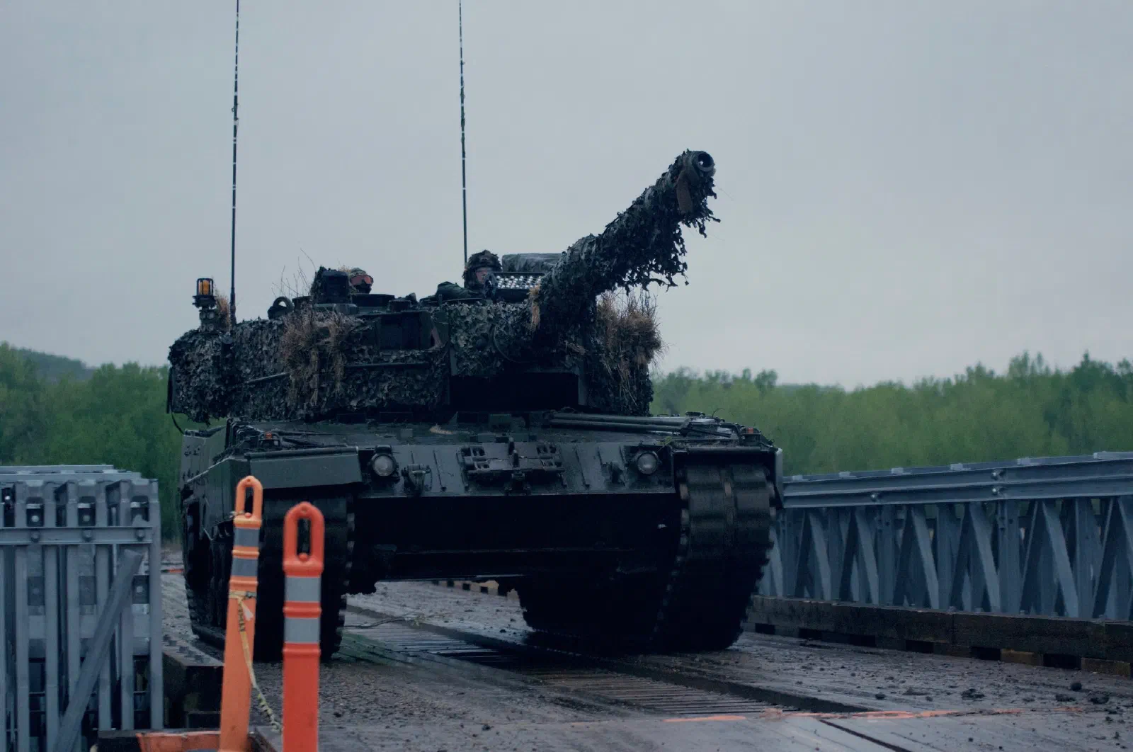 Өнгөлөн далдлах тор, өвсөөр бүрхсэн Канадын армийн Leopard 2A4 танк түр гүүрээр давж буй нь, урд нь улбар шар хаалтын шон, ар талд ногоон ой, бүрхэг тэнгэр