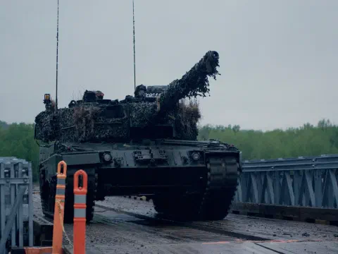 Өнгөлөн далдлах тор, өвсөөр бүрхсэн Канадын армийн Leopard 2A4 танк түр гүүрээр давж буй нь, урд нь улбар шар хаалтын шон, ар талд ногоон ой, бүрхэг тэнгэр